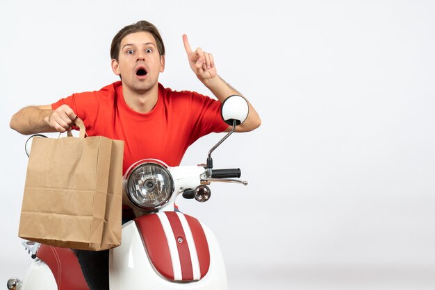 Young surprised courier guy in red uniform sitting on scooter holding paper bag and pointing up on yellow wall