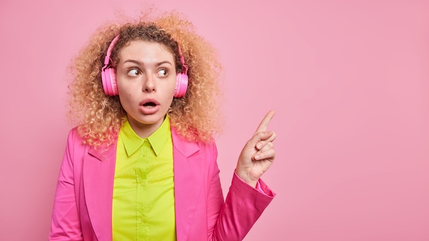 젊은 놀란 곱슬 머리 젊은 유럽 여자는 헤드폰을 통해 음악을 듣고 오른쪽 상단 모서리에 핑크 벽 위에 고립 된 공식적인 옷을 입고 있음을 나타냅니다.
