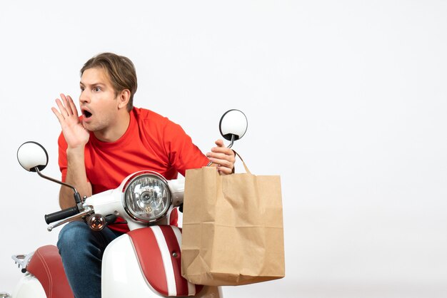 Young surprised delivery guy in red uniform sitting on scooter holding paper bag looking at somewhere on white wall