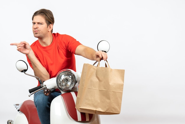Young surprised emotional courier guy in red uniform sitting on scooter giving paper bag pointing something on the right side on white wall
