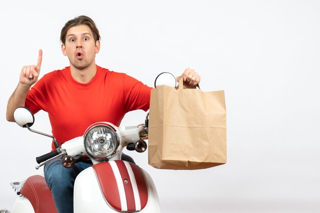 Young surprised emotional courier guy in red uniform sitting on scooter giving paper bag pointing up on white wall