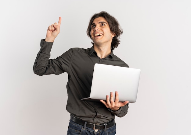 Молодой удивленный красивый кавказский мужчина держит ноутбук и указывает вверх на белом фоне с копией пространства
