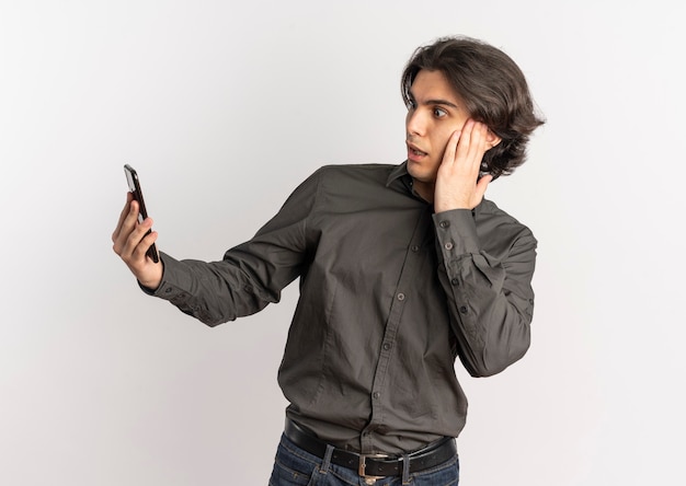 若い驚きのハンサムな白人男性は、コピースペースと白い背景で隔離の電話を保持し、見ています