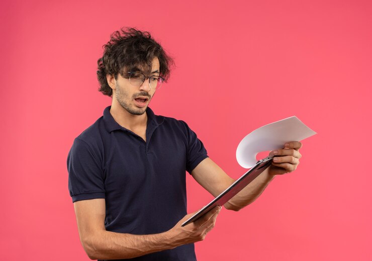 मुफ़्त फोटो young surprised man in black shirt with optical glasses holds and looks at clipboard isolated on pink wall