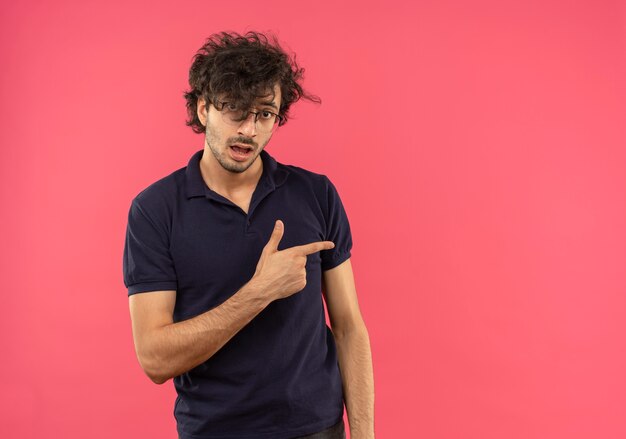 분홍색 벽에 고립 된 측면에서 광학 안경 포인트와 검은 셔츠에 젊은 놀란 남자