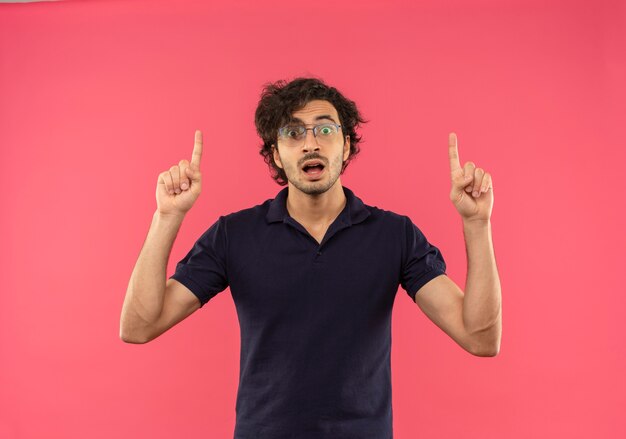 Young surprised man in black shirt with optical glasses points up and looks isolated on pink wall