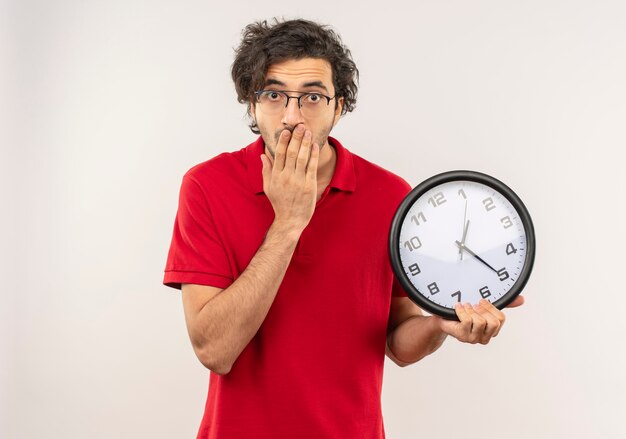 Young surprised man in red shirt with optical glasses holds clock and puts hand on mouth isolated on white wall