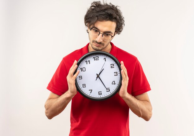 Young surprised man in red shirt with optical glasses holds and looks at clock isolated on white wall