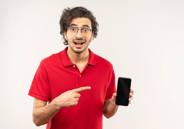 Young surprised man in red shirt with optical glasses holds and points at phone isolated on white wall
