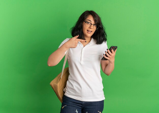 Young surprised pretty caucasian schoolgirl wearing glasses and back bag holds and points at phone on green  with copy space