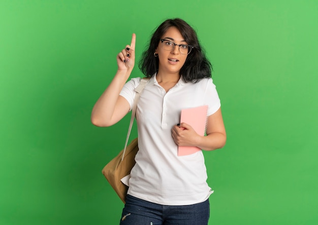 Young surprised pretty caucasian schoolgirl wearing glasses and back bag points up holding notebook on green  with copy space