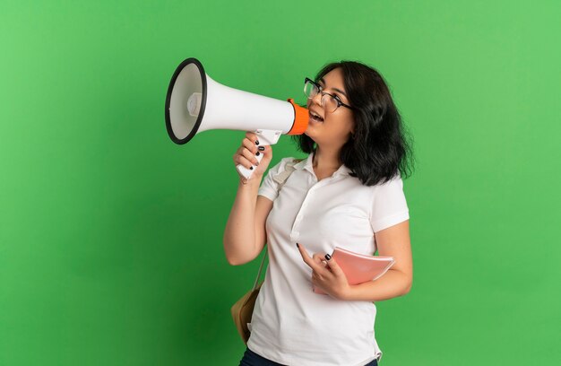 Young surprised pretty caucasian schoolgirl wearing glasses and back bag speaks through loud speaker holding notebook isolated on green space with copy space