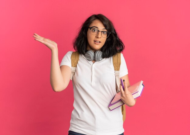 Young surprised pretty caucasian schoolgirl with headphones on neck wearing glasses and back bag holds books and points at side on pink  with copy space