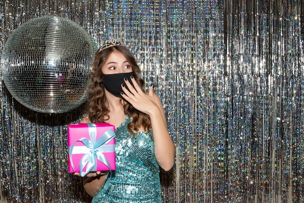 Young surprised pretty woman wearing shiny dress with sequins with crown in black medical mask and holding gift in the party