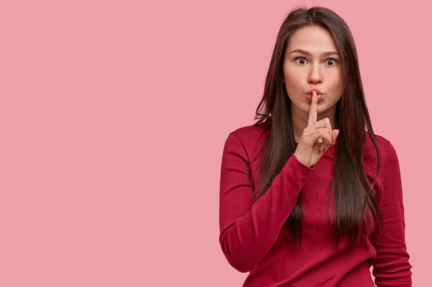 Young surprised woman makes silence gesture, looks with panicking expression, asks not tell secret, stares worried