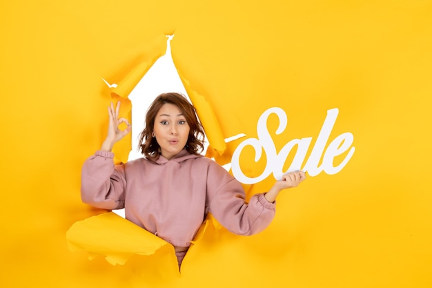 Free photo young surprised woman making eyeglasses gesture holding sale sign on yellow torn breakthrought background