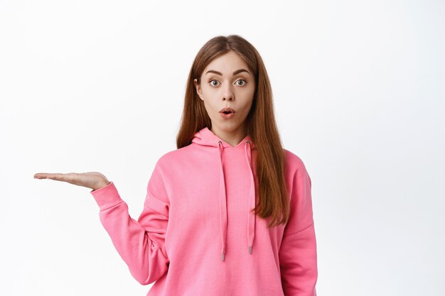 Young surprised woman say wow, holding empty space on palm, holding item on display, standing against white wall