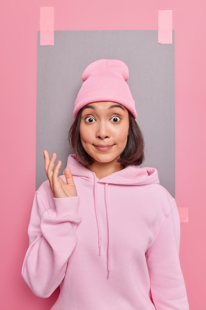 Young surprised young Asian woman stares bugged eyes keeps hand raised with indignation wears casual sweatshirt and hat poses against pink backgroud with blank empty space for your advertisement