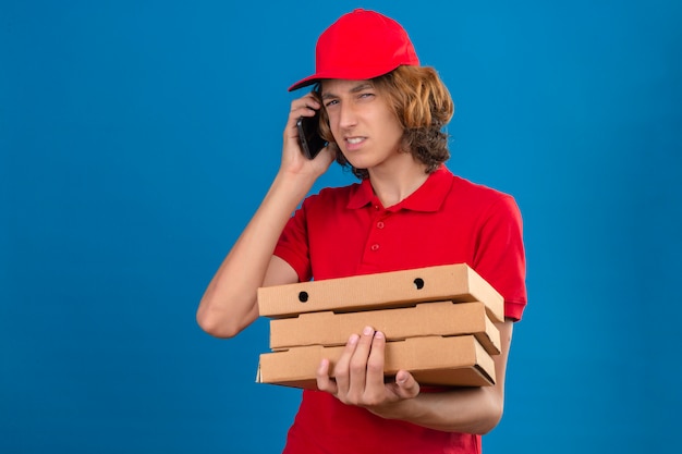 Young suspicious delivery man in red uniform talking on mobile phone while holding pizza boxes over isolated blue background