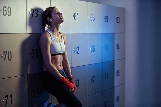 Молодая потная спортивная женщина отдыхает в раздевалке спортзала после спортивной тренировки и слушает музыку в наушниках