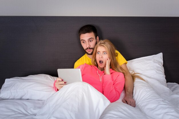 タブレットガジェットで何かを見ているベッドの上の若い甘いカップル。テクノロジーと人についての概念