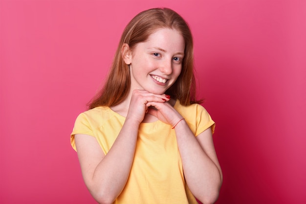 young sweet cute girl with smile on her face, holding arms near chin. Red haired lady with red manicure poses isolated on pink. People and emotions concept.