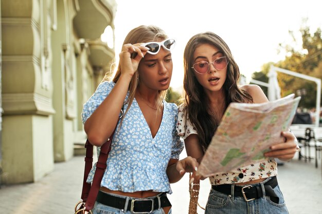 Young tanned blonde and brunette girls in sunglasses look at map outside Attractive tourists in floral blouses walk in city