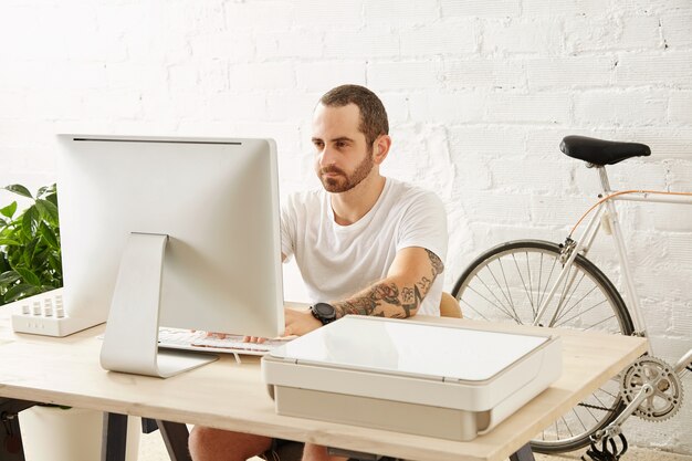 young tattooed freelancer man in blank white t-shirt works on his computer at home near his bicycle, looking in display