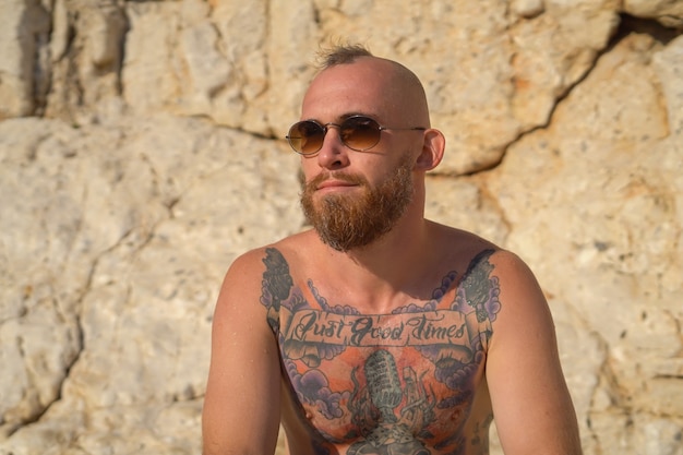 Free photo young tattooed sporty man on the beach on a hot summer sunny day in the mountain posing in sunglasses