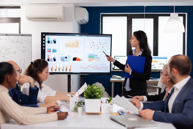 young team leader in big corporation briefing coworkers pointing at graph meeting corporate staff