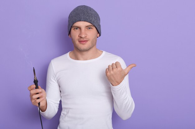 ワイヤーをはんだ付けする準備ができている若い技術者、魅力的な男性は白いカジュアルシャツを着て、灰色の帽子は片手ではんだごてを保持し、紫に分離された別の親指で脇を指します。