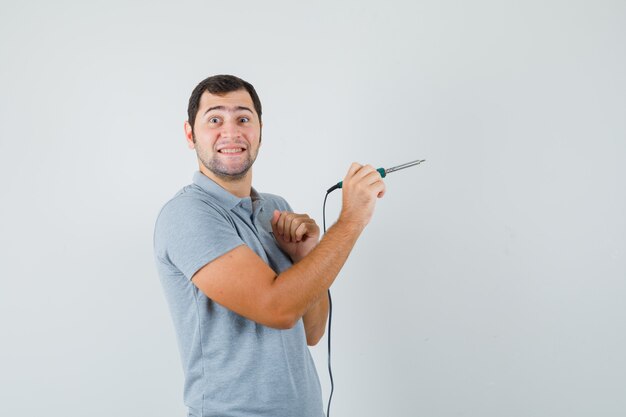 Молодой техник в серой форме держит дрель в одной руке, показывая большой палец вверх и выглядит веселым.