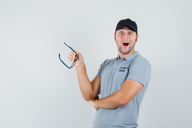 Young technician in grey uniform holding glasses and looking amazed , front view.