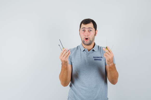 Young technician in grey uniform holding screwdrivers and looking surprised