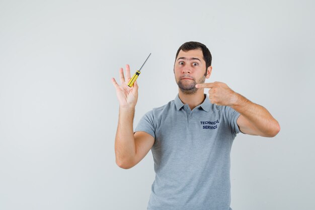 Young technician in grey uniform holding screwdrivers in one hand and pointing to it and looking serious.