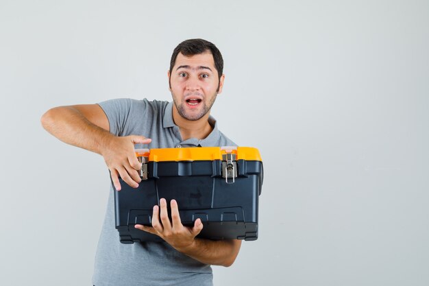 Young technician in grey uniform holding toolbox with his both hands and looking shocked.