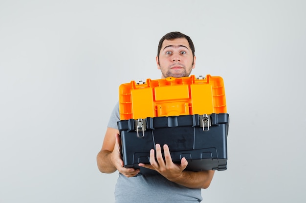 Young technician in grey uniform holding toolbox with his both hands and looking surprised.