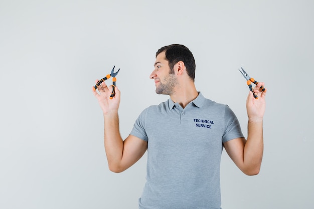 Young technician in grey uniform looking at the pliers holding in both hands and looking optimistic , front view.