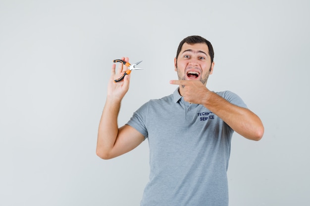 Young technician in grey uniform pointing at pliers and looking joyful , front view.