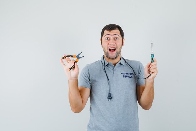 Young technician holding drill in one hand, plier in another hand in grey uniform and looking dazed. front view.