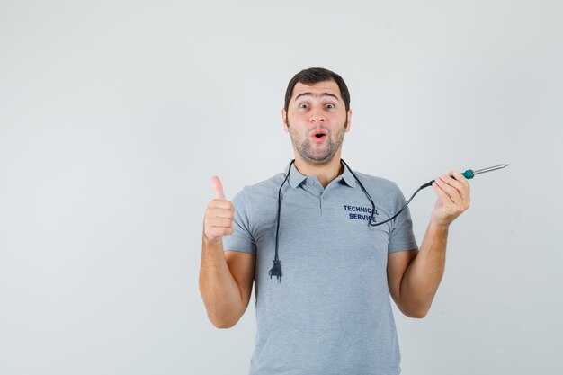 Young technician holding drill in one hand while showing thumb up in grey uniform and looking surprised.