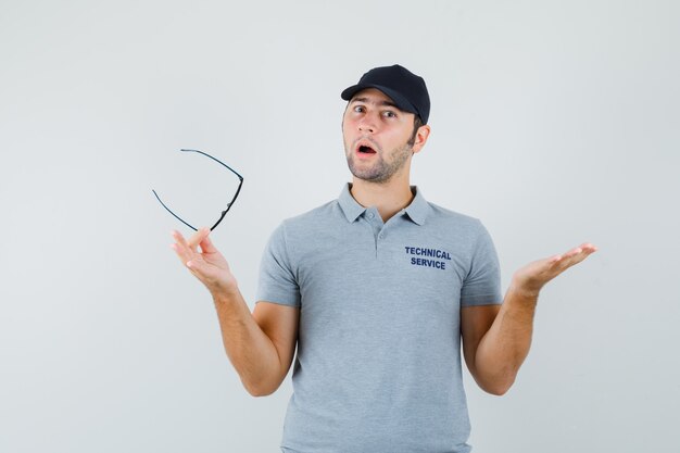 Young technician holding glasses in grey uniform and looking confused.