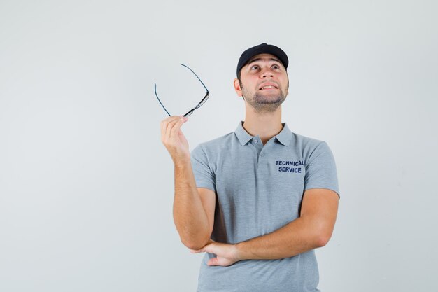 Young technician holding glasses in grey uniform and looking thoughtful.