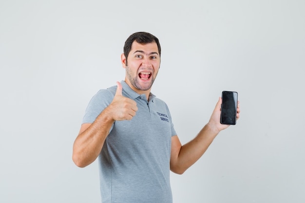 Young technician holding mobile phone, showing thumb up in grey uniform and looking blissful , front view.