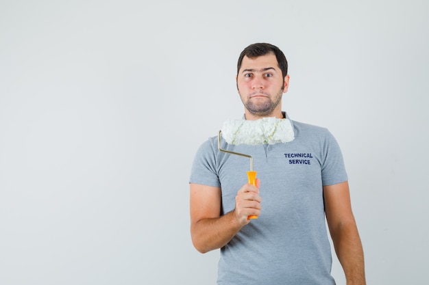 Foto gratuita giovane tecnico che tiene il rullo di vernice in uniforme grigia e che sembra serio.