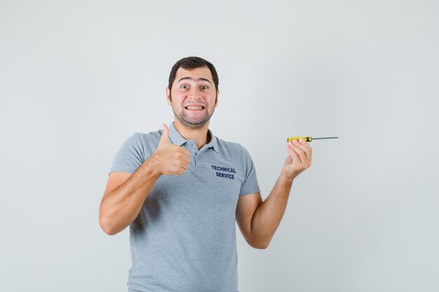 Young technician holding screwdriver, showing thumb up in grey uniform and looking joyful.