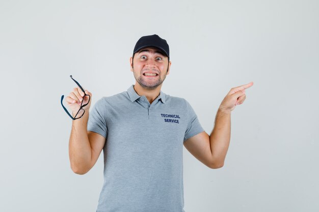 Young technician pointing aside, holding glasses in grey uniform and looking jolly.