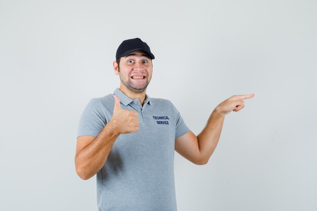 Young technician pointing aside, showing thumb up in grey uniform and looking glad.