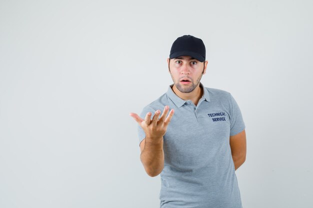 Young technician stretching hand in puzzled gesture in grey uniform front view.