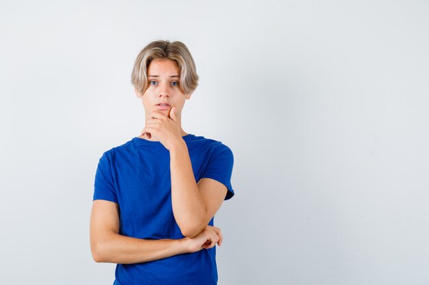 Young teen boy in blue t-shirt with hand on chin and looking dismal , front view.